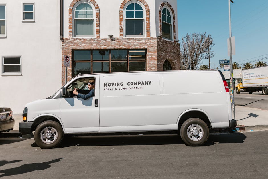 A white moving van with the text 'MOVING COMPANY LOCAL & LONG DISTANCE' printed on the side is parked on a street in front of a multi-storey brick building with large glass windows. The van is positio