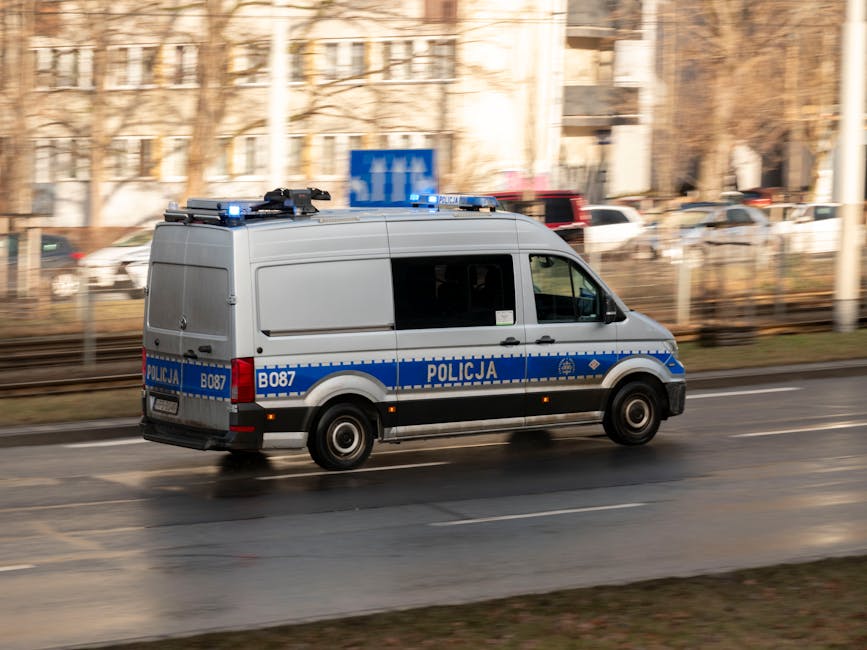 A white police van with blue and black markings, labeled 'POLICJA' and with vehicle identification B087, is moving along a wet urban street. The van is equipped with a roof-mounted light bar and is photographed in motion, causing a slight blur effect. The surrounding environment includes a railway track fence, multiple parked cars, and a multi-storey building in the background, with leafless trees indicating winter. The wet road surface reflects the van’s movement, and the overall scene appears to be during daylight hours. The image exemplifies the type of vehicle that may be encountered during city relocations or when law enforcement assistance is involved in moving or security logistics, with Man With a Van White City providing comprehensive moving services, including transportation and associated logistics.