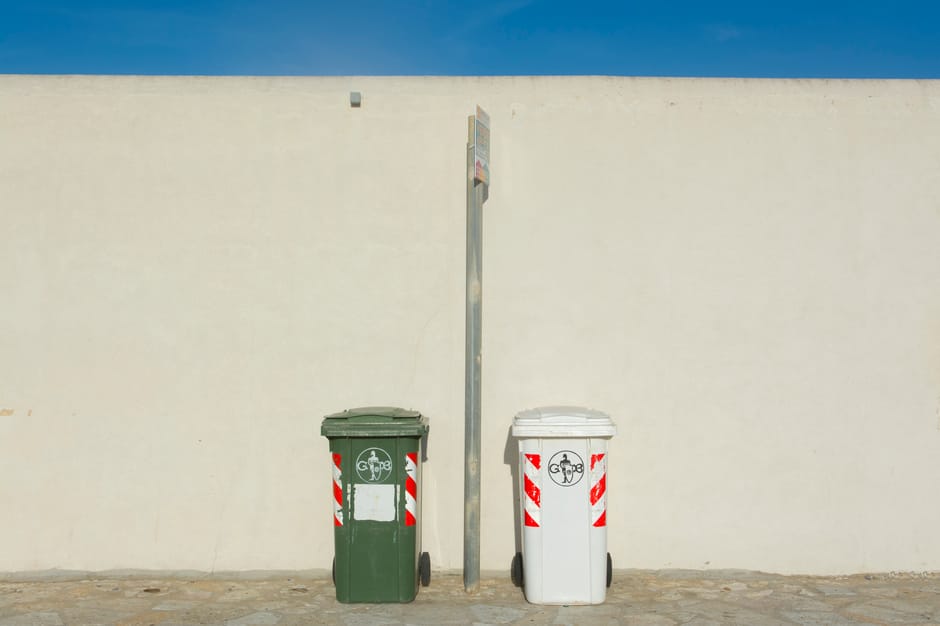 A photograph depicting two wheeled waste bins positioned on a paved area in front of a plain, off-white exterior wall under bright daylight. The bin on the left is green with a closed lid, and the bin on the right is white, also with a closed lid. Both bins are marked with recycling symbols and red-and-white striped reflective tape on their sides, indicating waste segregation. Between the bins, there is a tall, slender metal pole with a small, rectangular sign attached at the top, which appears to be a bus stop sign. The setting suggests a residential or commercial property exterior, and the scene may relate to household waste collection during a home relocation or moving process, illustrating waste disposal practices in accordance with controlled waste laws in White City. Man With a Van White City, a professional removals service, may use such images to demonstrate proper waste management during house moves.