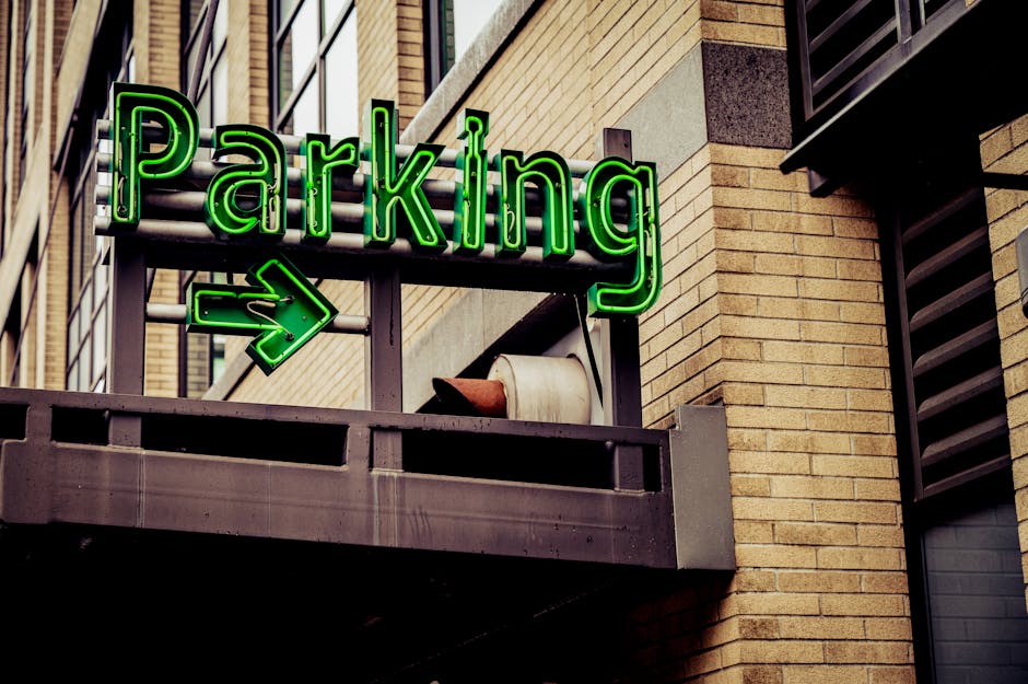 A close-up view of a balcony sign that reads 'Parking' in bright green neon lights, with a green arrow pointing to the left beneath the word. The sign is mounted on a dark metal frame attached to the exterior brick facade of a modern building, which features a mixture of light and dark brown bricks. An empty white plastic plant pot rests on the balcony railing beside the sign. The surrounding environment includes windows with black frames and louvered vents, indicating an urban setting. This scene highlights the importance of understanding parking regulations for home relocation and furniture transport, as evidenced by the prominent signage that might be relevant for house removals services provided by Man With a Van White City, especially when planning loading and unloading logistics around restricted parking areas.