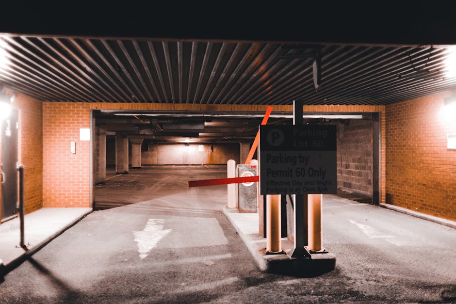 The image shows the entrance to an underground parking garage with a brick wall exterior and a dark, low ceiling with metal slats. In the foreground, a small sign indicating parking restrictions is mounted on a concrete barrier, and the barrier is equipped with red and orange safety poles. The driveway leading into the garage is paved with asphalt and has white directional arrows painted on it. Inside, the parking area appears empty, with concrete pillars supporting the structure and low lighting illuminating the space. On the left side at the entrance, a metal gate is partially visible. This setting is typical for vehicle loading during home relocation or furniture transport, where parking permits and regulation compliance are essential. The presence of the sign and barriers suggests the need for parking permits or adherence to local parking rules for moving services, as provided by Man With a Van White City, for efficient house removals and logistics in the White City area.