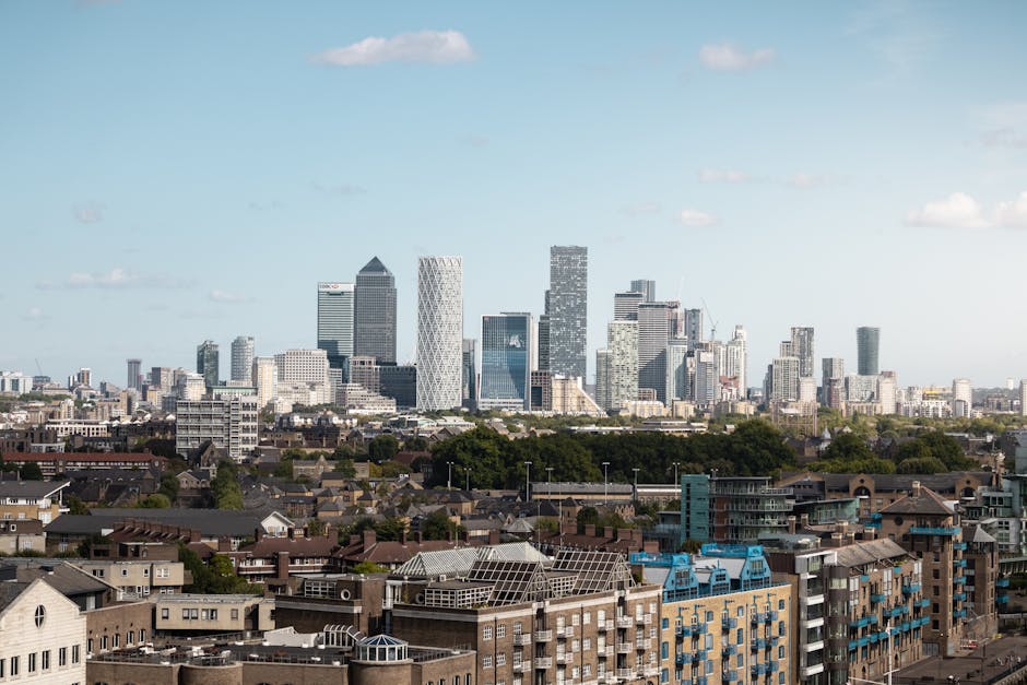 A panoramic view of the White City skyline with modern high-rise office buildings and skyscrapers in the background under a blue sky with scattered clouds, while in the foreground, there are lower residential and commercial buildings with flat and pitched roofs, some with solar panels. The image captures the urban environment during daylight, showcasing the mix of residential housing and the central business district where valuable furnishings and equipment are typically transported during home relocation or moving processes. This scene reflects the kind of urban setting where professional removals services, such as those provided by Man With a Van White City, are often employed to facilitate home removals and furniture transport within the city, especially in busy commercial and residential zones.