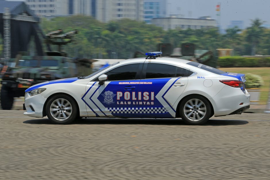 A silver police car with blue and white markings is shown in motion on a paved road, with the word 'POLISI' written prominently on the side, indicating it is a Malaysian police vehicle. The car is fully visible, including the blue light bar mounted on the roof, and the vehicle's windows are dark-tinted. In the background, there are blurred images of trees, greenery, and some distant buildings, suggesting the vehicle is moving through an urban or suburban area. The road surface appears to be asphalt, and the scene is captured during daylight hours with natural lighting. The police car is associated with law enforcement, and the activity seen is consistent with routine patrol or transportation, set against a backdrop that does not inform about specific surroundings or ongoing incidents.