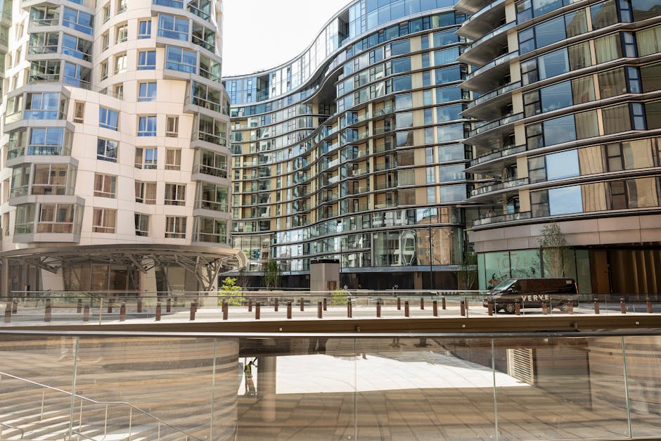 A panoramic view of the White City skyline with modern high-rise office buildings and skyscrapers in the background under a blue sky with scattered clouds, while in the foreground, there are lower residential and commercial buildings with flat and pitched roofs, some with solar panels. The image captures the urban environment during daylight, showcasing the mix of residential housing and the central business district where valuable furnishings and equipment are typically transported during home relocation or moving processes. This scene reflects the kind of urban setting where professional removals services, such as those provided by Man With a Van White City, are often employed to facilitate home removals and furniture transport within the city, especially in busy commercial and residential zones.