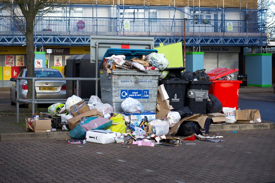 A large collection of overflowing waste and recycling bins positioned on a paved sidewalk in front of commercial premises located in Hammersmith & Fulham, with various types of rubbish including cardboard boxes, paper, plastic bags, and packaging materials scattered around. The waste containers include a blue bin designated for mixed paper and card, a black bin for general waste, and a red bin, all placed behind a metal barrier rail on a street corner. In the background, a blue van is partially visible, indicating a home relocation or moving process by Man With a Van White City, amidst ongoing street renovations visible with scaffolding and building signage. The scene captures the typical logistical challenges faced during household moves and the importance of proper waste disposal in accordance with local regulations, especially relevant for residents or movers involved with the updated bulky waste rules governed by Hammersmith & Fulham Council.