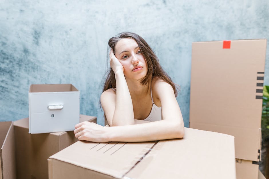 A young woman with long brown hair and a neutral expression rests her head on her hand while sitting among several cardboard moving boxes of varying sizes, some sealed with packing tape. The boxes are arranged on the floor and on a flat surface in front of her, indicating a packing or unpacking process in a home environment, possibly during a house move. Behind her is a plain, light blue wall, and to her right, a larger cardboard box stands upright, with a smaller, white storage box placed on one of the other boxes. The scene is well-lit with natural light, suggesting daytime, and the setting appears to be an indoor space prepared for a home relocation. The presence of packing materials and boxes reflects typical packing and moving activities associated with professional removals, such as those provided by Man With a Van White City, especially in relation to compliance with local waste disposal guidelines like the Hammersmith & Fulham Council bulky waste rules.