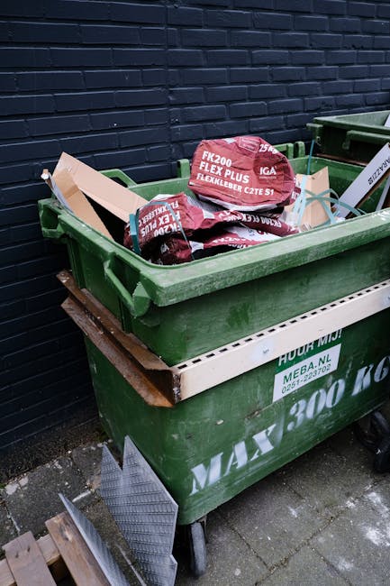 A large collection of overflowing waste and recycling bins positioned on a paved sidewalk in front of commercial premises located in Hammersmith & Fulham, with various types of rubbish including cardboard boxes, paper, plastic bags, and packaging materials scattered around. The waste containers include a blue bin designated for mixed paper and card, a black bin for general waste, and a red bin, all placed behind a metal barrier rail on a street corner. In the background, a blue van is partially visible, indicating a home relocation or moving process by Man With a Van White City, amidst ongoing street renovations visible with scaffolding and building signage. The scene captures the typical logistical challenges faced during household moves and the importance of proper waste disposal in accordance with local regulations, especially relevant for residents or movers involved with the updated bulky waste rules governed by Hammersmith & Fulham Council.