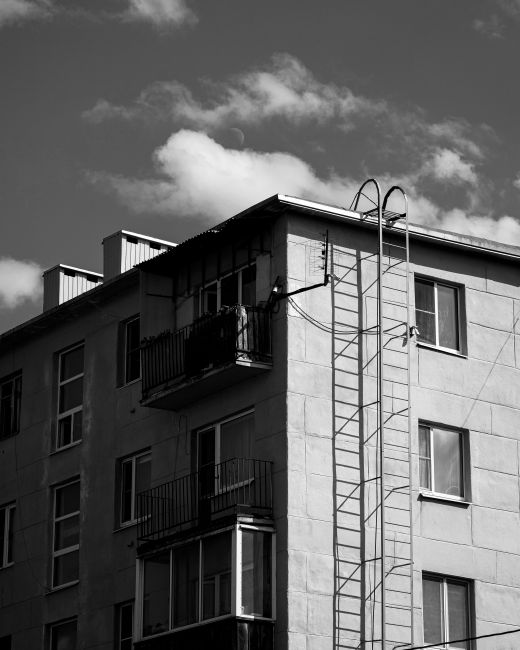 A black-and-white photograph of a multi-storey residential building with several balconies, some of which have metal railings and awnings. The building's exterior is composed of large concrete or ston