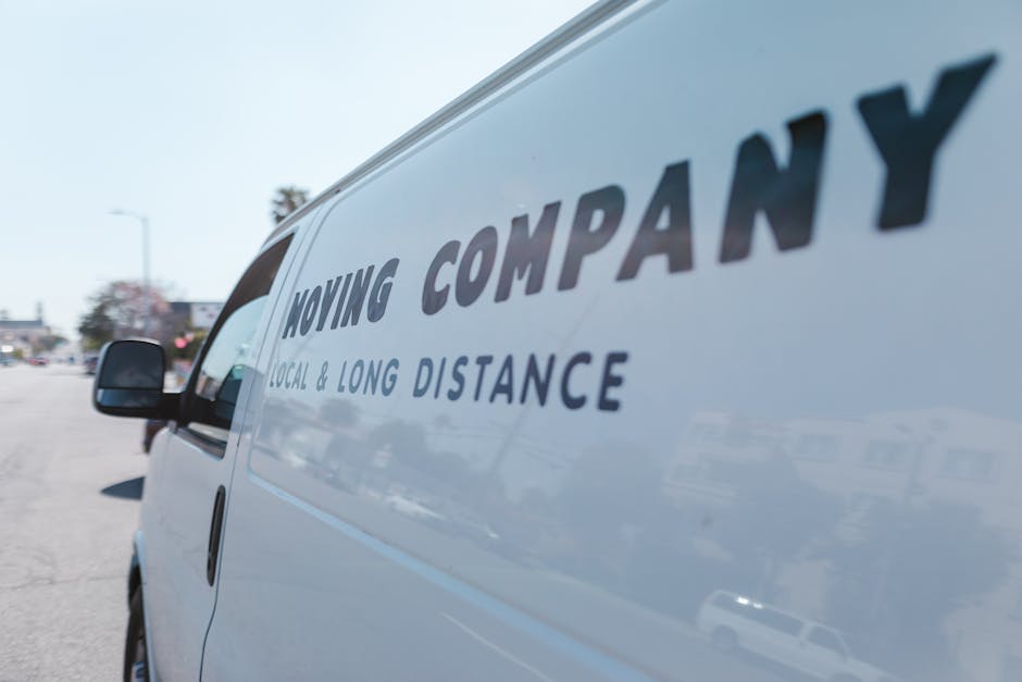 A close-up view of a white moving van parked outside a residential property during daylight hours, displaying the text 'MOVING COMPANY' in large, bold letters along with 'LOCAL & LONG DISTANCE' beneath it. The van's side door is visible, with a side mirror and part of the vehicle's window in view. In the background, there are other parked cars, a few trees, and a clear sky, suggesting a suburban environment. The scene appears to be part of a home relocation or furniture transport process, with the van ready for loading or recently unloaded. This image reflects the professional movement services provided by Man With a Van White City as part of their removals offering.