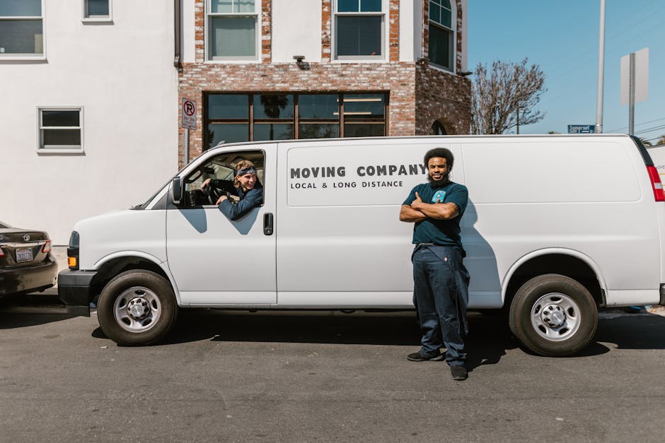 A smiling young man with long hair and a bandana around his head is sitting in the driver’s seat of a white moving van with the company name 'Moving Company' and the words 'Local & Long Distance' printed on the side. The van is parked outside a residential building with brick walls, large windows, and a clear blue sky overhead. Inside the van’s open door, a variety of packed cardboard boxes, wrapped furniture with plastic or fabric covers, and packing materials can be seen arranged for a home relocation. The man is dressed casually in a dark shirt, appearing ready for furniture transport and loading. The van is positioned on the pavement, indicating it is during a house move, and the image captures the loading process involved in professional removals using a moving vehicle. [COMPANY_NAME] is involved in coordinating the packing, transport, and logistics of the house removals, reflecting efficient moving services.