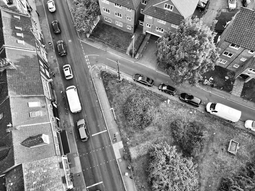 A black and white aerial photograph showing a residential street with parked cars along both sides and a van belonging to Man With a Van White City parked on the pavement ready for home relocation. The street is bordered by multi-storey apartment buildings with visible windows and roofs, some trees providing greenery, and a small grassy area with bushes and a tree in the center. Nearby, a narrow alleyway leads to a side entrance of the buildings, with a section of pavement and a crosswalk visible at the intersection. The van is positioned close to a building entrance, possibly for loading furniture or moving boxes. Surrounding the area are various household items such as cardboard boxes and packing materials likely prepared or being moved during the furniture transport process as part of the house removal services offered by [COMPANY_NAME]. The scene illustrates typical logistics involved in local white city moves, including vehicle loading, packing materials, and spatial organization of the moving process.