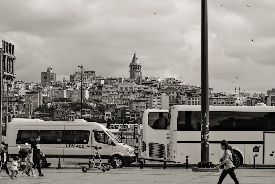 A black and white image shows a street scene with two white passenger vans and a bus parked on the roadside, likely used for home relocation or furniture transport. The vans are positioned side by side with their rear doors closed, and the bus is parked behind them. In the foreground, a man wearing a dark T-shirt and face mask is walking past a family of three, including a young child holding a small bicycle, and a woman is walking in the opposite direction. The scene takes place on a paved pavement with short metal bollards and a lamppost nearby. In the background, a densely built hillside cityscape features iconic architecture, including the Galata Tower, under a cloudy sky. The setting, with active loading and unloading of furniture and boxes, indicates a professional removal or moving service, likely managed by Man With a Van White City, supporting local house moves such as from Latimer Road to Shepherd's Bush.
