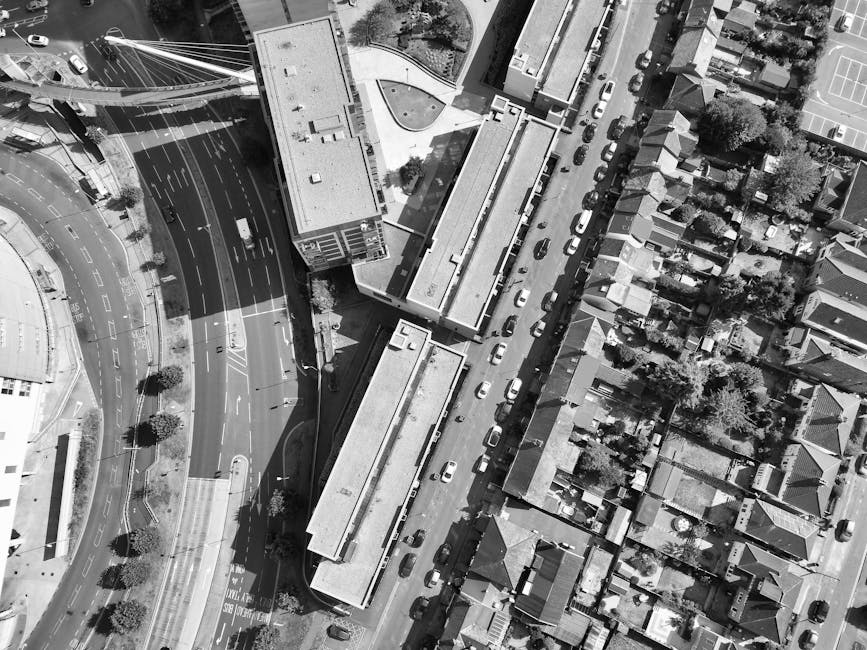 A black and white aerial photograph showing a residential street with parked cars along both sides and a van belonging to Man With a Van White City parked on the pavement ready for home relocation. The street is bordered by multi-storey apartment buildings with visible windows and roofs, some trees providing greenery, and a small grassy area with bushes and a tree in the center. Nearby, a narrow alleyway leads to a side entrance of the buildings, with a section of pavement and a crosswalk visible at the intersection. The van is positioned close to a building entrance, possibly for loading furniture or moving boxes. Surrounding the area are various household items such as cardboard boxes and packing materials likely prepared or being moved during the furniture transport process as part of the house removal services offered by [COMPANY_NAME]. The scene illustrates typical logistics involved in local white city moves, including vehicle loading, packing materials, and spatial organization of the moving process.