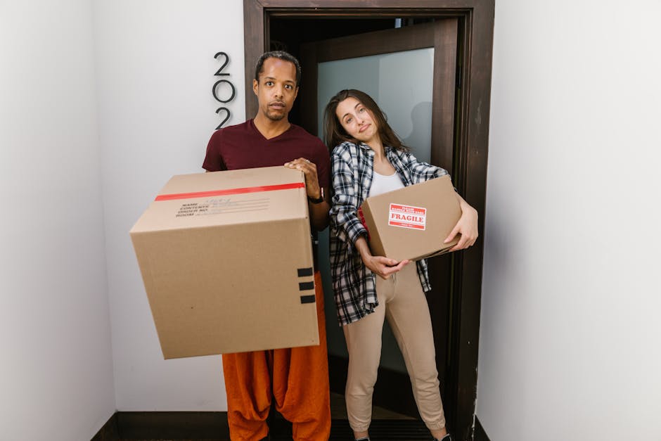 A man and a woman stand inside a doorway, each holding a cardboard box as part of a home relocation process. The man, positioned on the left, wears a maroon t-shirt and orange trousers, holding a large cardboard box sealed with red and black packing tape. The woman, on the right, wears a plaid shirt over a white top and beige trousers, carrying a smaller box marked with a red 'Fragile' label. They appear to be preparing for moving or loading furniture and boxes for transport, with the doorway leading to an interior space and the door slightly open behind them. The scene suggests an organized moving process, possibly coordinated by Man With a Van White City, as part of comprehensive packing and furniture transport services related to a house removal or relocation in Westfield London, specifically White City.