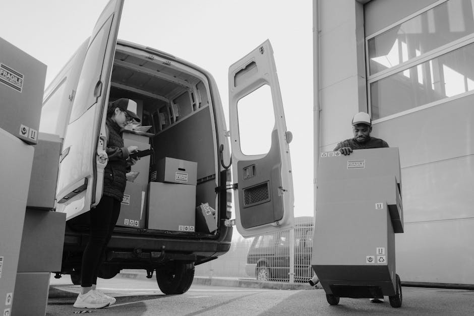 Two removal workers from Man With a Van White City are engaged in the loading process outside a commercial property, with the back doors of a large white moving van open. The female worker, dressed in casual clothing including a cap and jacket, is inside the van organizing or checking boxes, which are made of cardboard and vary in size, some sealed with packing labels. She is holding a clipboard or tablet. The male worker, wearing a cap and a safety helmet, is outside the van, lifting or placing a large cardboard box onto a flat trolley or dolly. Several other boxes are positioned nearby on the pavement, ready for transport. The scene takes place during daylight, with the surrounding environment including a modern building with large windows and a metal fence, indicating an urban setting suitable for home or office relocations. The setup reflects professional furniture transport and packing elements integral to a move or removals service, as provided by Man With a Van White City.
