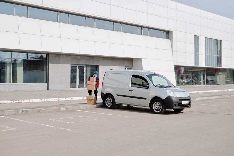 A person wearing a blue beanie and red jacket is standing next to a white delivery van parked in an outdoor, paved parking area in front of a modern building with large glass windows and white cladding. The individual is carrying several cardboard boxes, some stacked on the ground and others in their hands, indicating a home relocation or moving process. The van is positioned close to the building's entrance, with its sliding side door closed. The scene is well-lit with natural daylight, and the parking lot appears to be mostly empty, with marked parking spaces visible. This setting illustrates a typical loading and unloading phase during furniture transport or packing and moving activities associated with house removals, which is relevant to services offered by Man With a Van White City, especially in the context of navigating permit issues on Wood Lane for efficient logistics.
