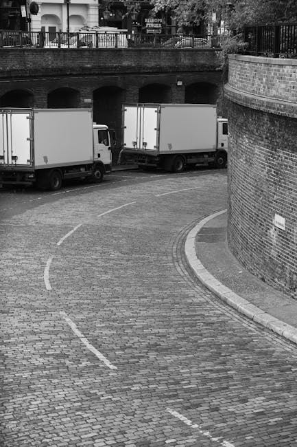 Two removal workers from Man With a Van White City are engaged in the loading process outside a commercial property, with the back doors of a large white moving van open. The female worker, dressed in casual clothing including a cap and jacket, is inside the van organizing or checking boxes, which are made of cardboard and vary in size, some sealed with packing labels. She is holding a clipboard or tablet. The male worker, wearing a cap and a safety helmet, is outside the van, lifting or placing a large cardboard box onto a flat trolley or dolly. Several other boxes are positioned nearby on the pavement, ready for transport. The scene takes place during daylight, with the surrounding environment including a modern building with large windows and a metal fence, indicating an urban setting suitable for home or office relocations. The setup reflects professional furniture transport and packing elements integral to a move or removals service, as provided by Man With a Van White City.