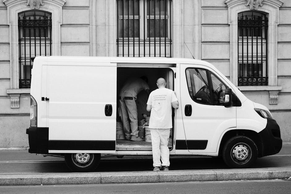 A white cargo van parked on a residential street in White City, with the background featuring an old, multi-story apartment building with weathered concrete walls and multiple windows, some of which h