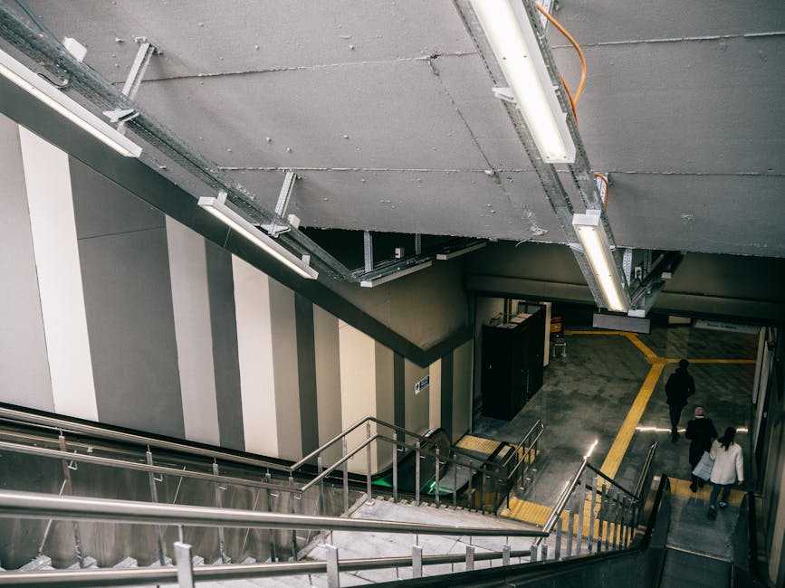 The image shows an indoor staircase and escalator area within a multi-story building, featuring striped wall designs in grey and white. The staircase has metal handrails and steps, with some areas covered by protective padding or blankets, indicating a moving or handling process. Two individuals are walking along the lower level, one carrying a bag and the other holding a small item, while a third person stands nearby, all dressed in dark and light clothing. The ceiling above has exposed structural elements and lighting fixtures, with visible electrical wiring and conduit. The environment appears to be in a commercial or residential property, with a paved floor section marked by yellow lines suggesting a designated area for moving or loading. This scene depicts a professional move facilitated by [COMPANY_NAME], focusing on efficient furniture transport, packing, and logistical planning in an accessible interior space, suitable for home relocation or apartment moves.