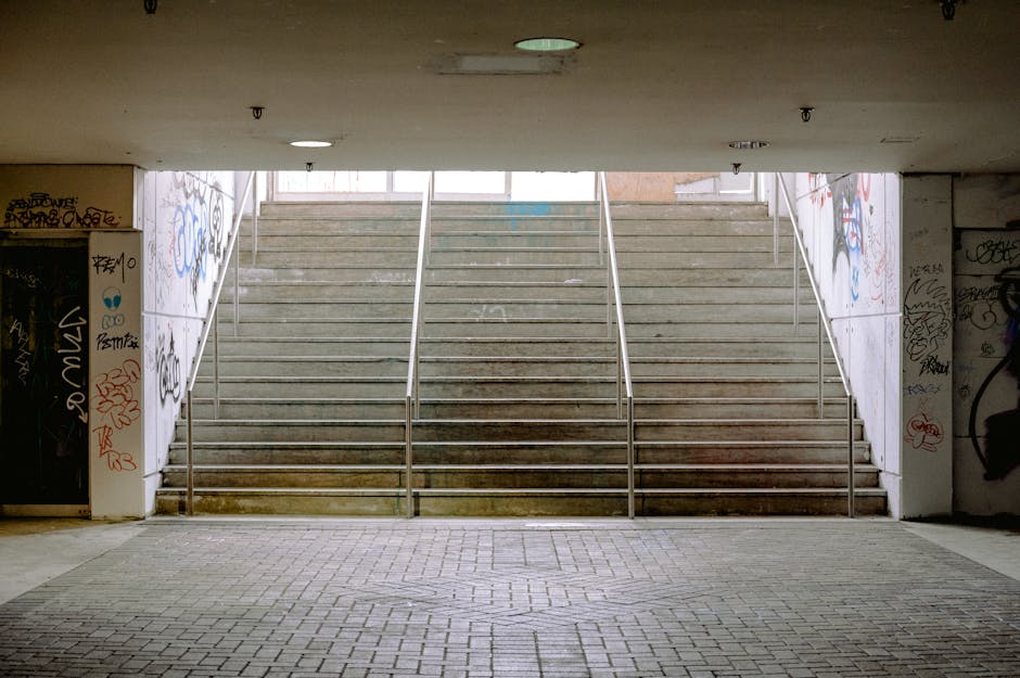 The image depicts an underground stairwell leading up to the street level at White City estate, visible through an open passageway under a concrete bridge or overpass. The stairs are made of concrete and are divided into three sections by metal handrails, with each section featuring a series of steps leading upwards. The walls on either side of the staircase are covered in graffiti, with various tags and artwork in different colors. The floor at the base of the stairs is paved with rectangular bricks, creating a level paved area suitable for loading and unloading. The underside of the overpass is plain, with a few ceiling lights providing illumination. The overall scene suggests a typical urban access point for residents or movers, where loading/unloading activities may take place as part of house relocation services provided by Man With a Van White City, particularly relevant to stairs, lifts, and access considerations during moving processes.